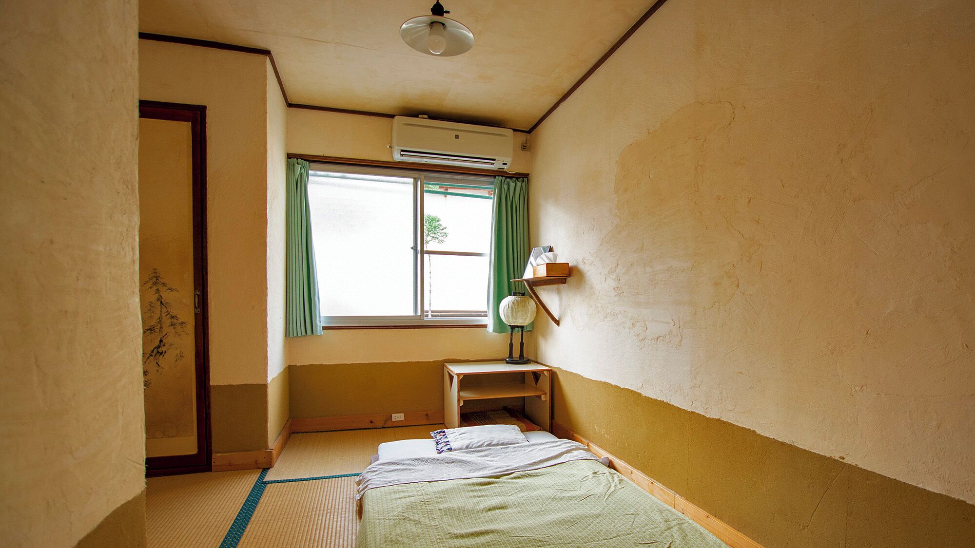 Habitació amb terra de tatami, un llit, una taula petita amb una làmpada, una estantalla amb llibres i un aparell d'aire condicionat.