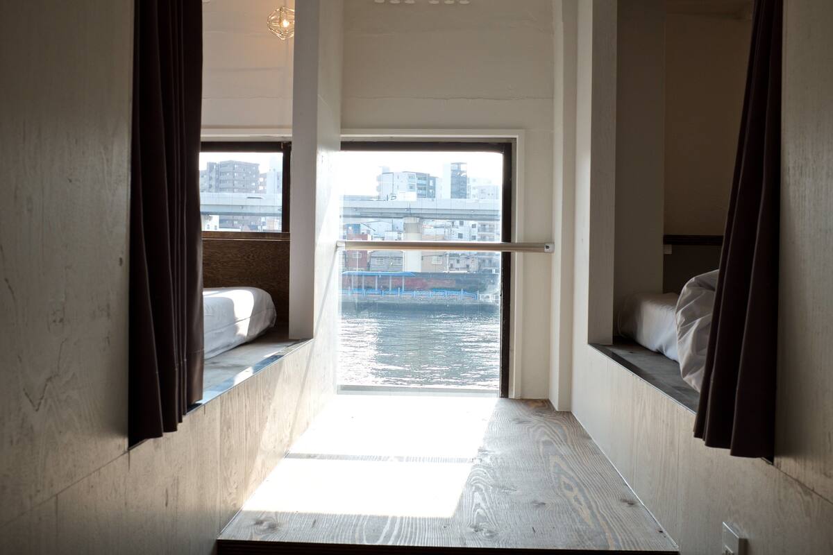 Habitación estrecha con vistas a una vía acuática y edificios. Hay dos camas con cortinas.