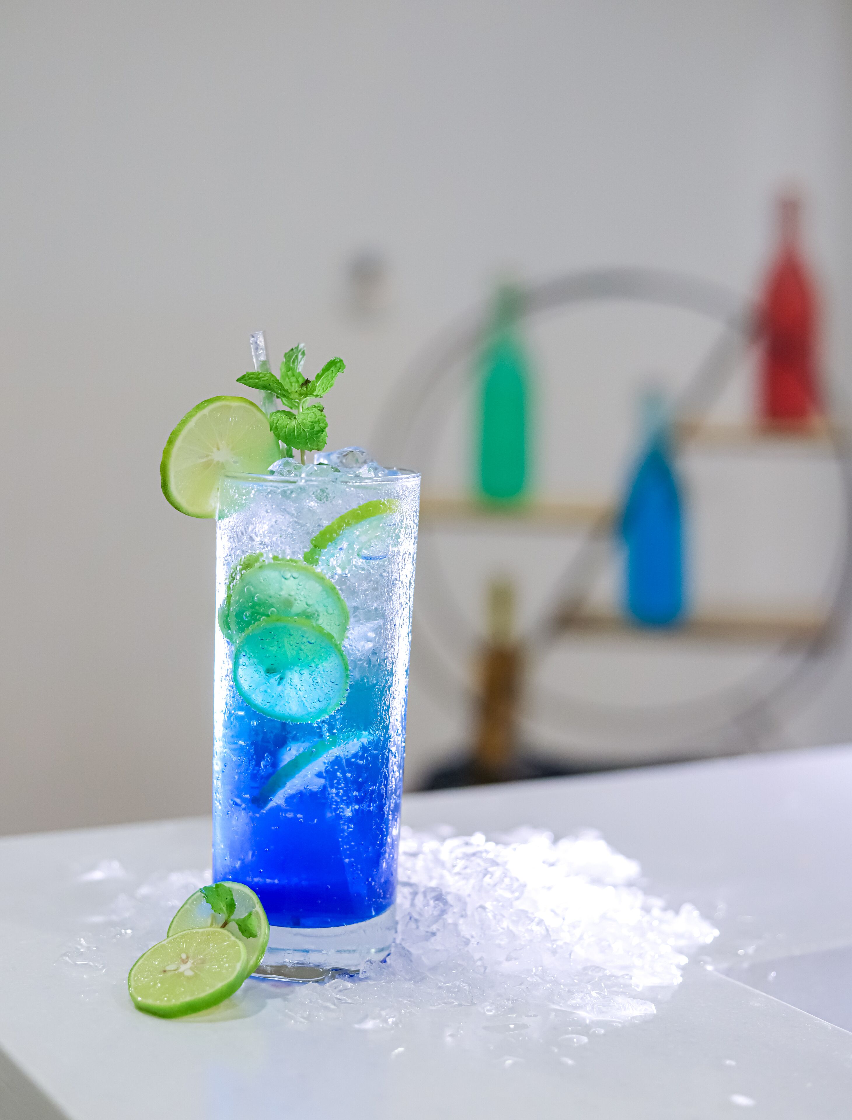 A tall glass of blue beverage with lime slices and mint garnish on a white surface with ice.