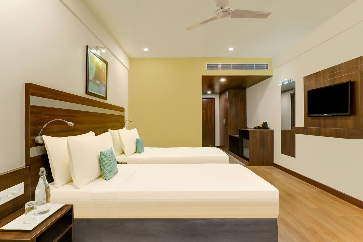 Une chambre d’hôtel avec deux lits, une télévision, un ventilateur de plafond et une salle de bain visible à travers une porte ouverte.