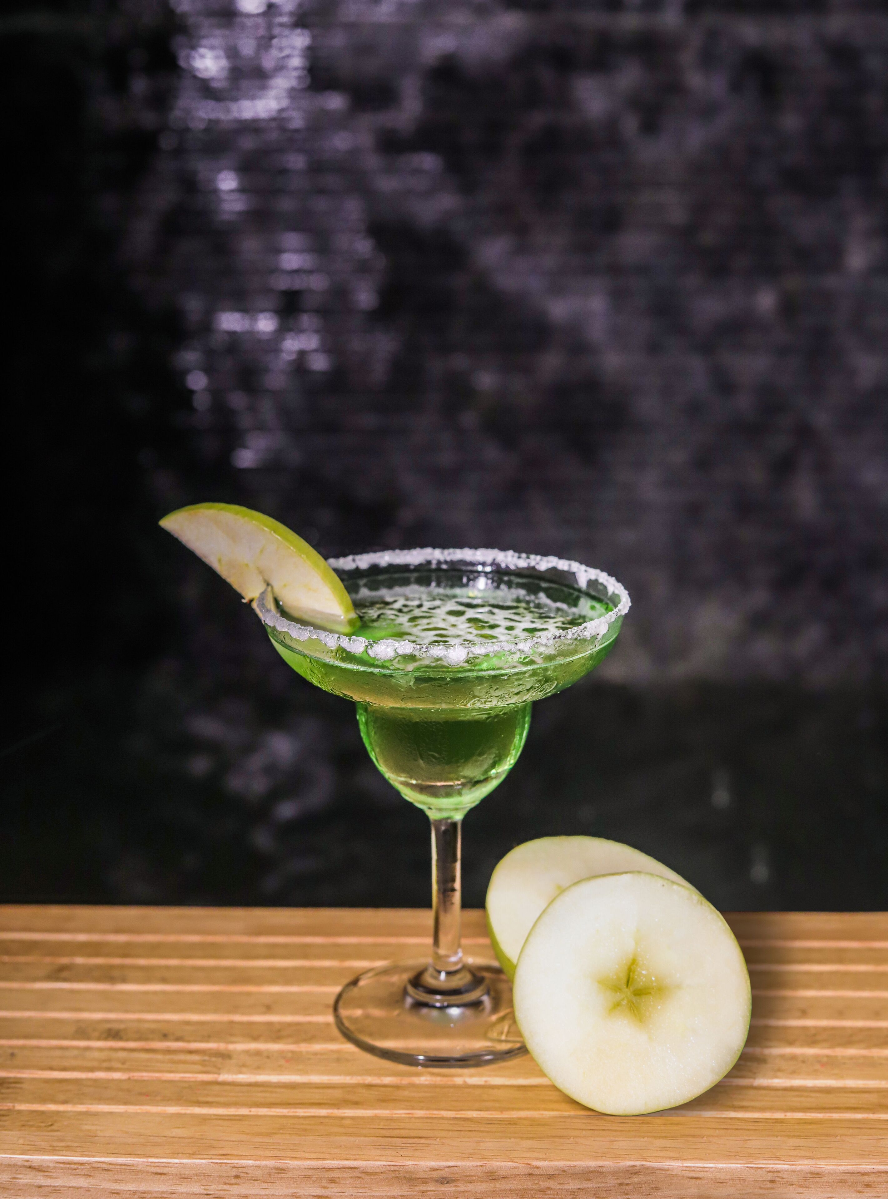 A green cocktail in a martini glass with a lime garnish, accompanied by a halved apple on a wooden surface.