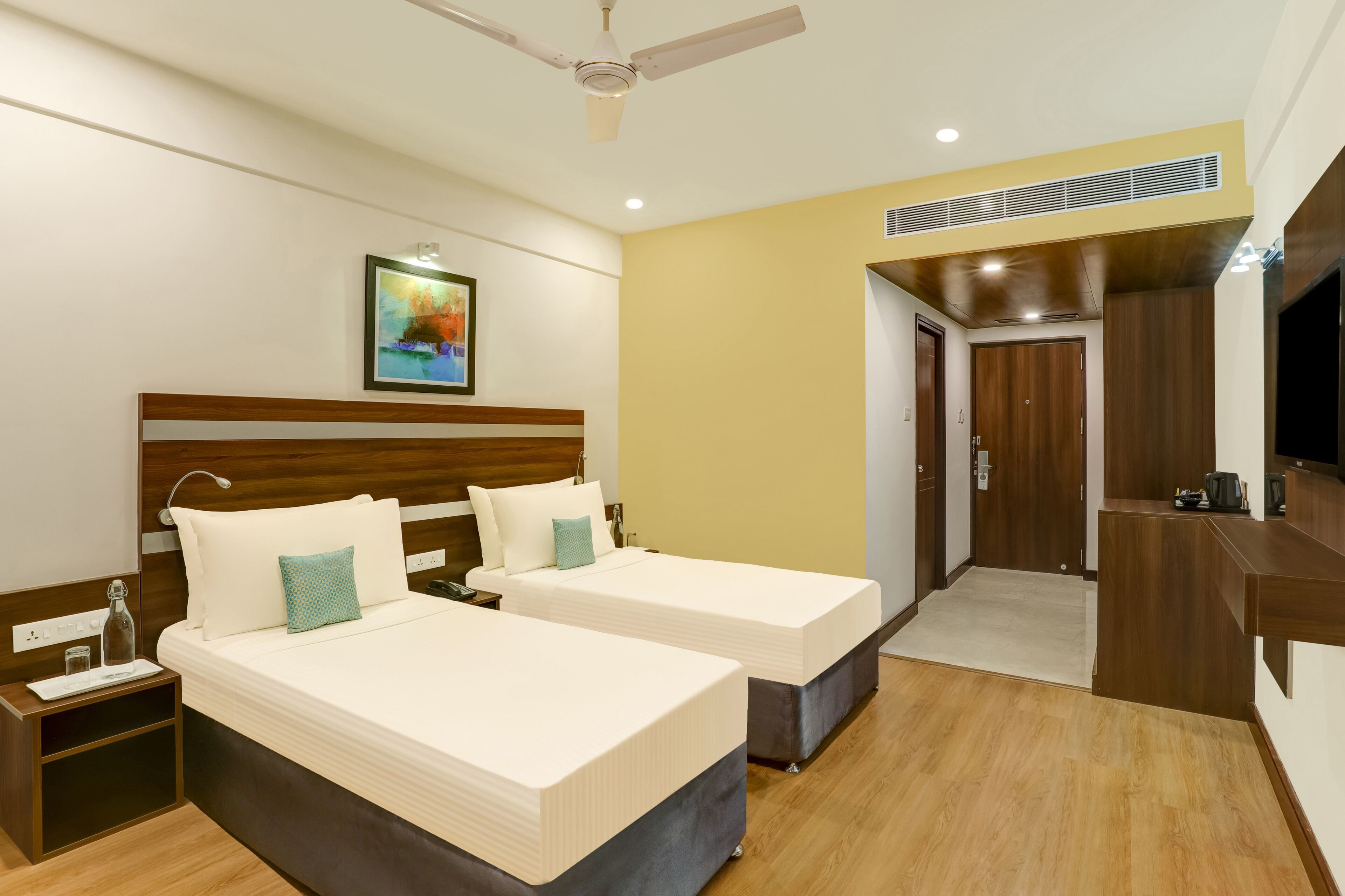 A hotel room with two beds, a wooden headboard, a ceiling fan, a painting, and a wooden desk with a chair.