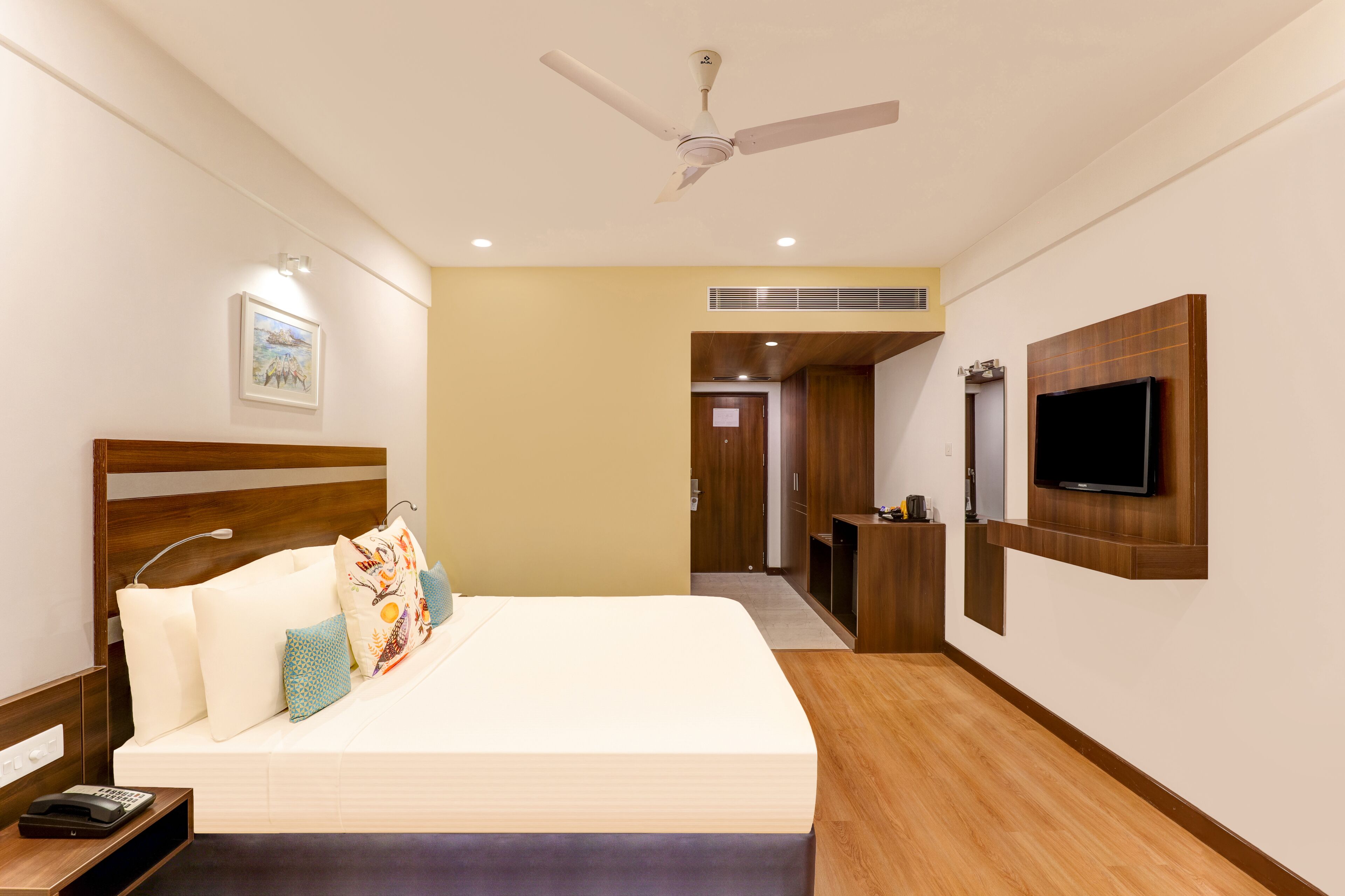 A hotel room with a bed, a TV mounted on the wall, a ceiling fan, and a wooden floor.