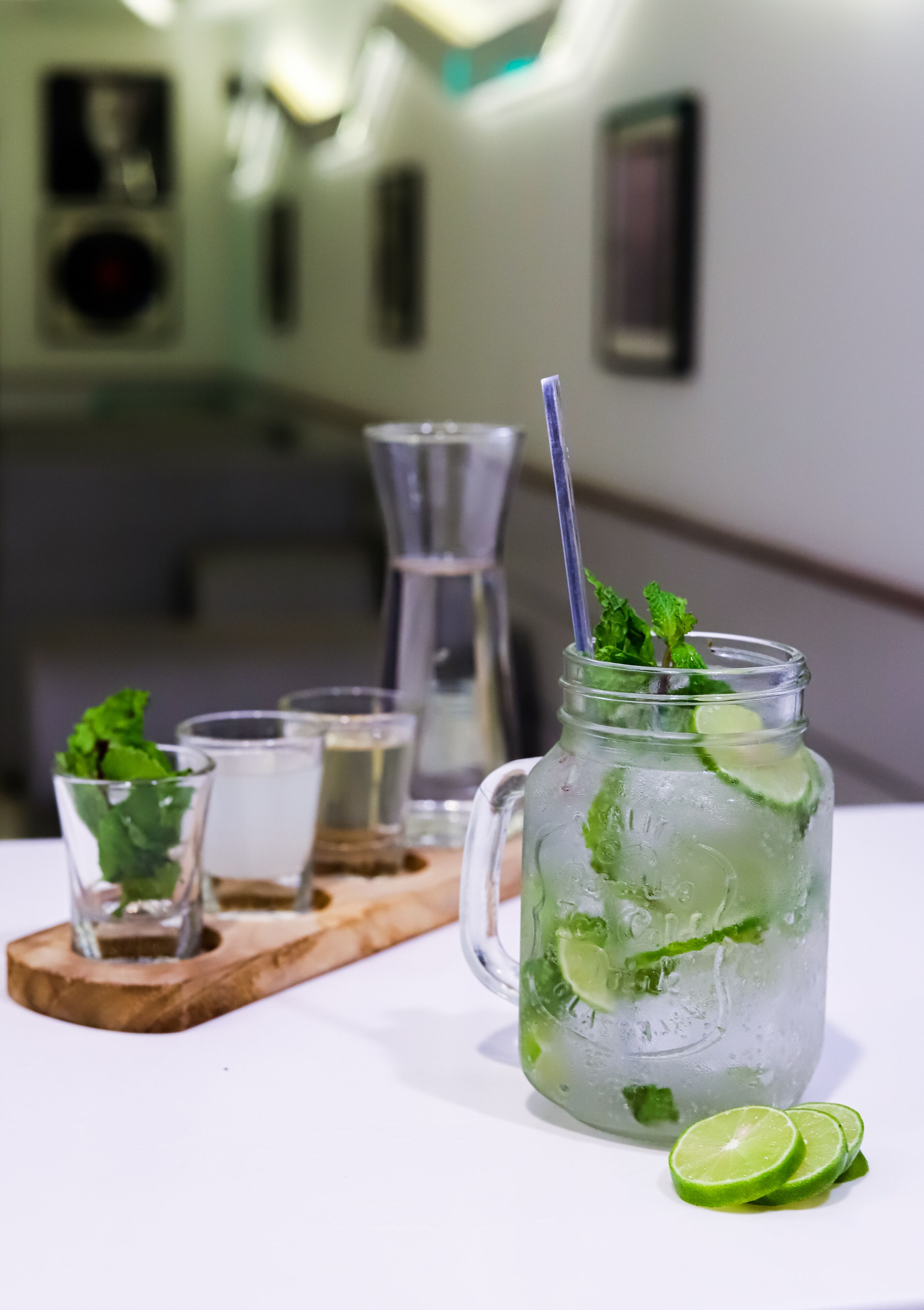 A glass of iced beverage with lime and mint, accompanied by two shot glasses and a wooden tray.