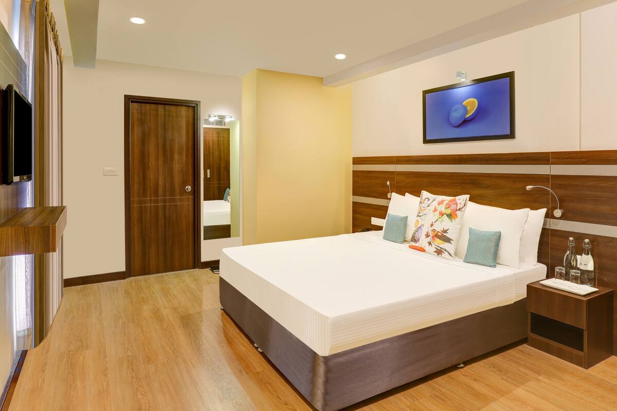 Une chambre d’hôtel moderne avec un grand lit, une tête de lit en bois, un téléviseur à écran plat et une petite table de chevet avec une bouteille et un verre.