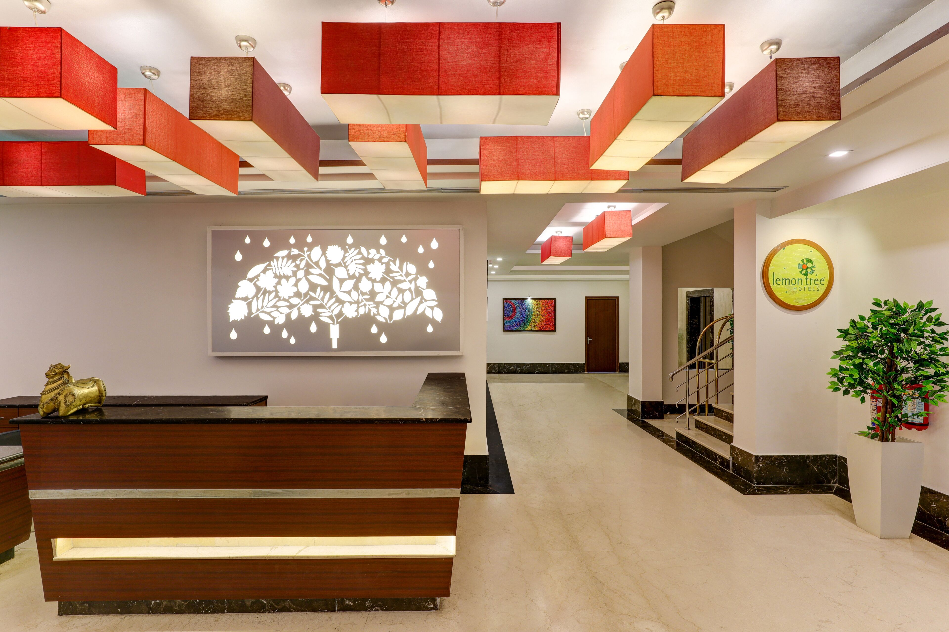 Un hall d’hôtel avec un comptoir d’accueil en bois, un luminaire décoratif en forme de fleurs fixé au mur, une peinture abstraite aux couleurs vives et un panneau indiquant « lemontree hotels ».