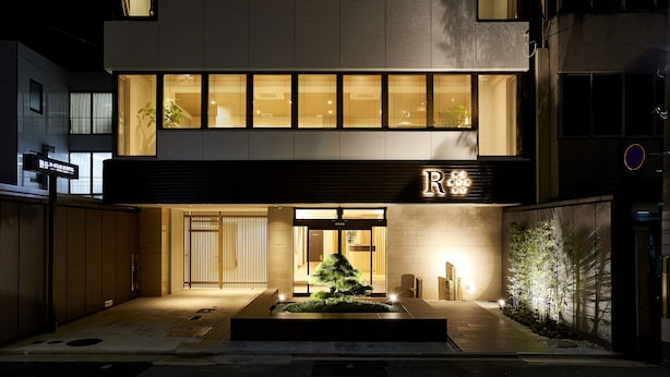 A modern hotel entrance with a well-lit facade, a reception desk, and a small landscaped area with a tree.
