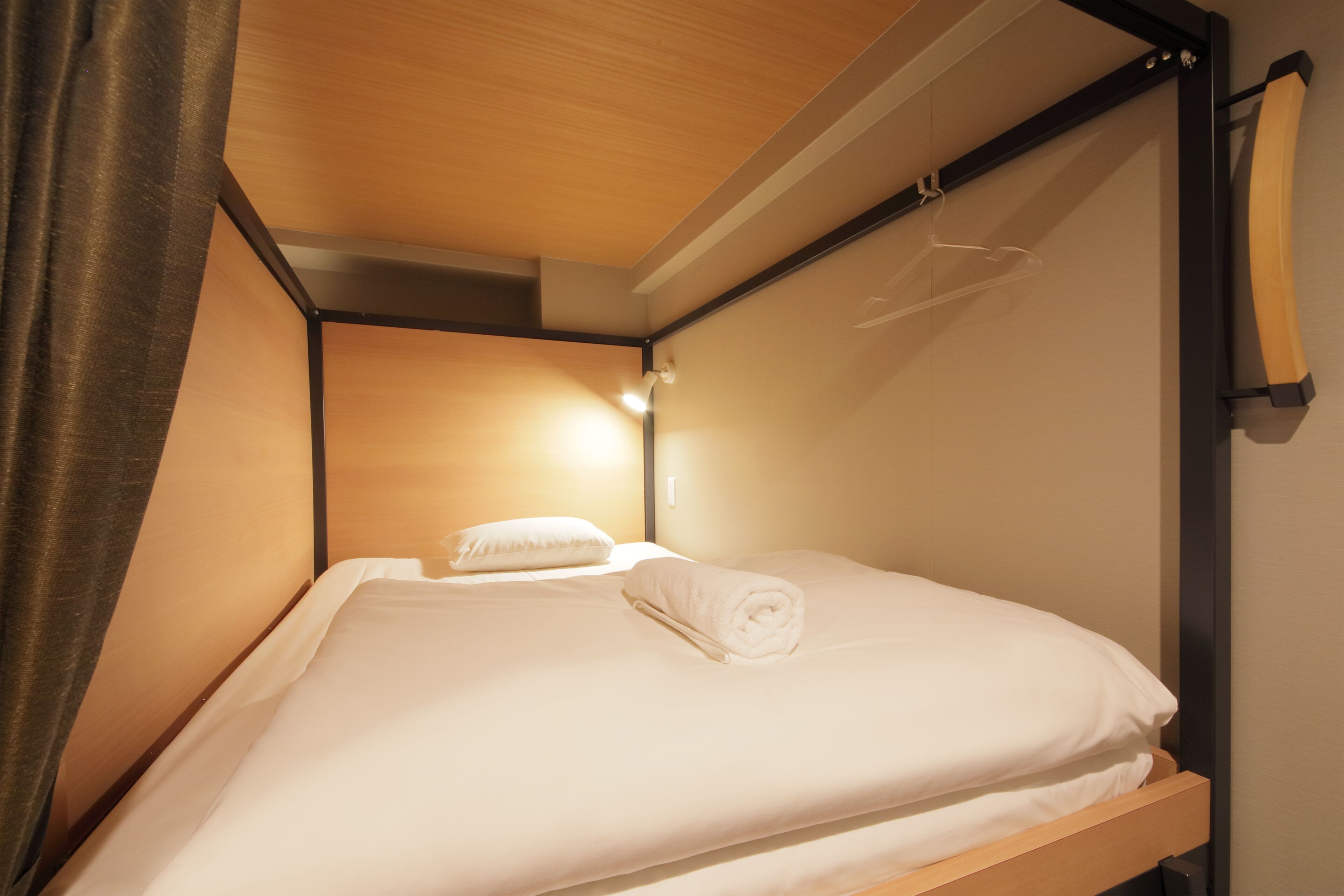 A single bed with a wooden frame and white bedding, a hanging light fixture, and a curtain partially covering the window.