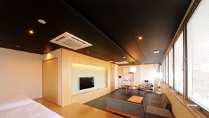 A modern hotel room with a wooden floor, a sofa, a TV, and a dining area with a table and chairs.