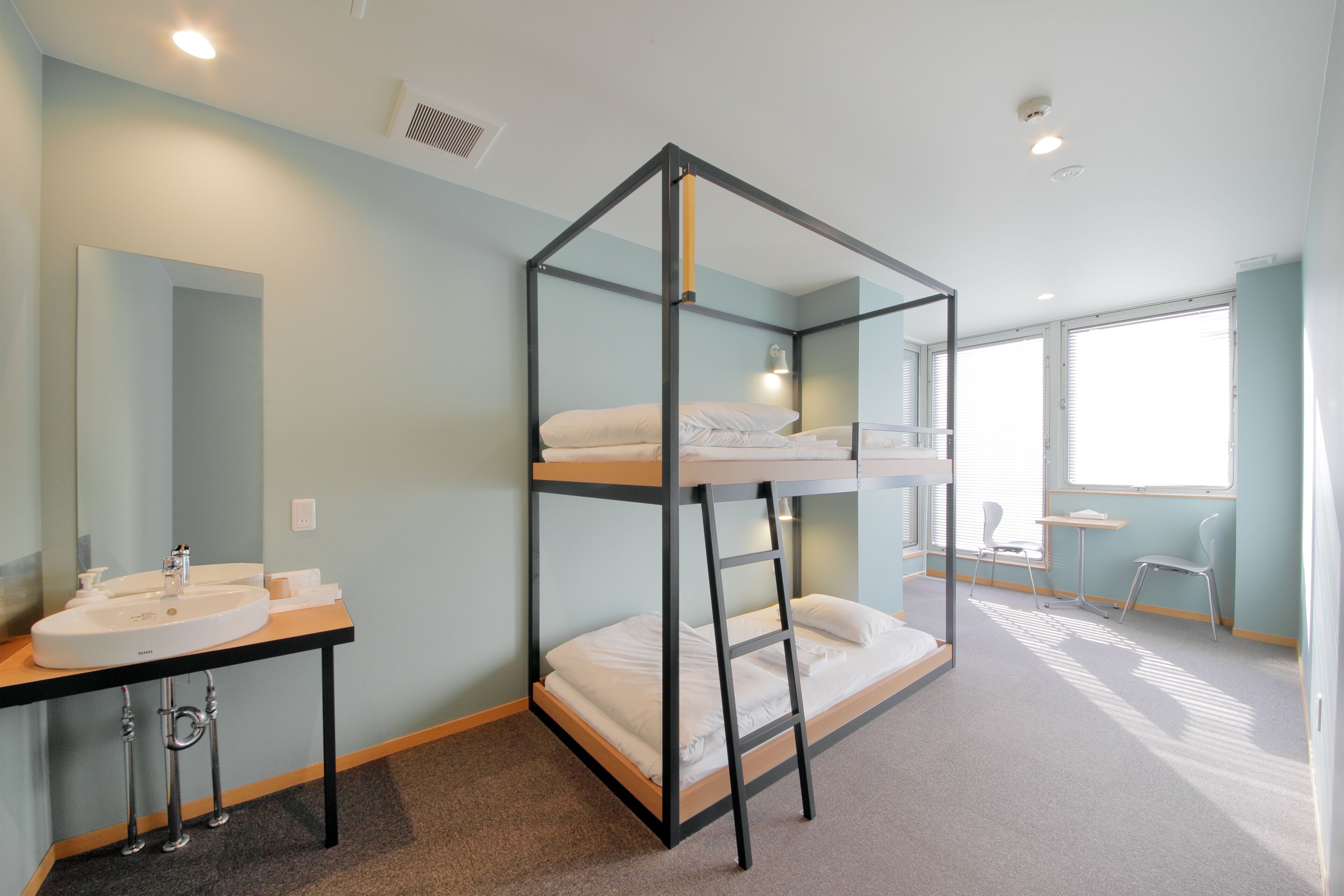A bunk bed with a desk and chair, a sink, and a mirror in a room with light blue walls and large windows.