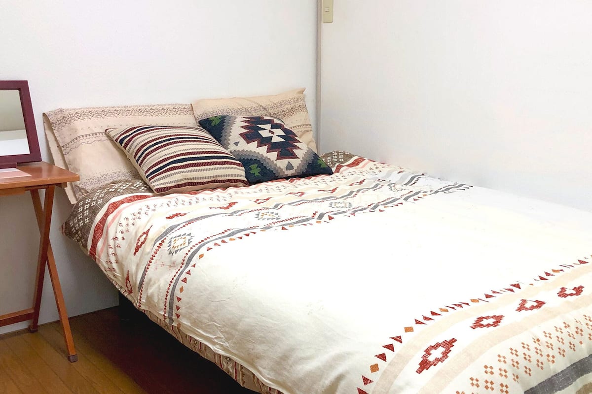 A small, neatly made bed with patterned bedding and pillows, a wooden nightstand with a mirror, and a plain white wall.