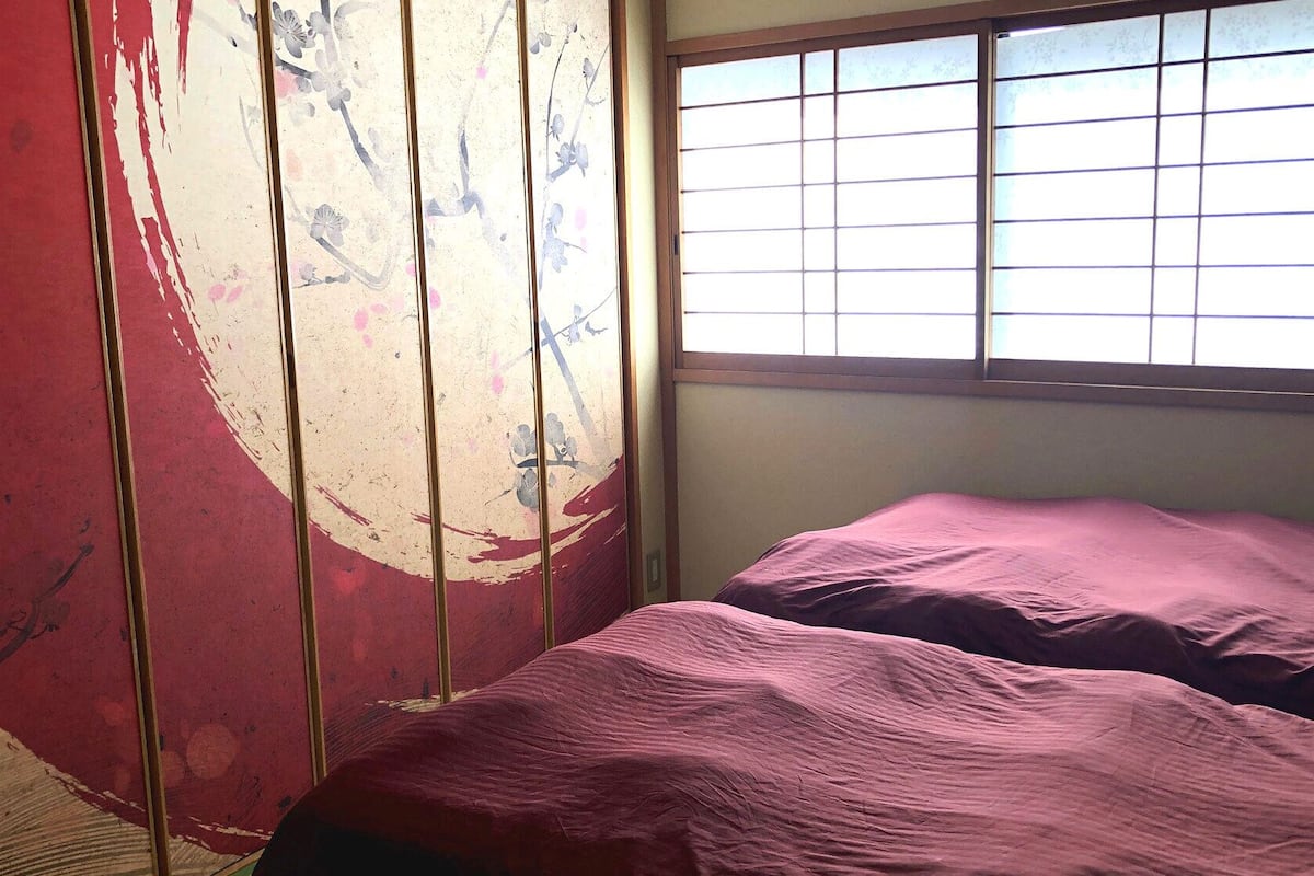 A room with a bed, a window with bars, and a wall with a partial red and white design.