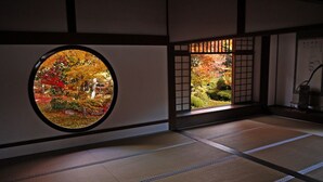 Una habitación japonesa tradicional con suelo de tatami, un celosía de papel y una ventana redonda con vistas al follaje otoñal.
