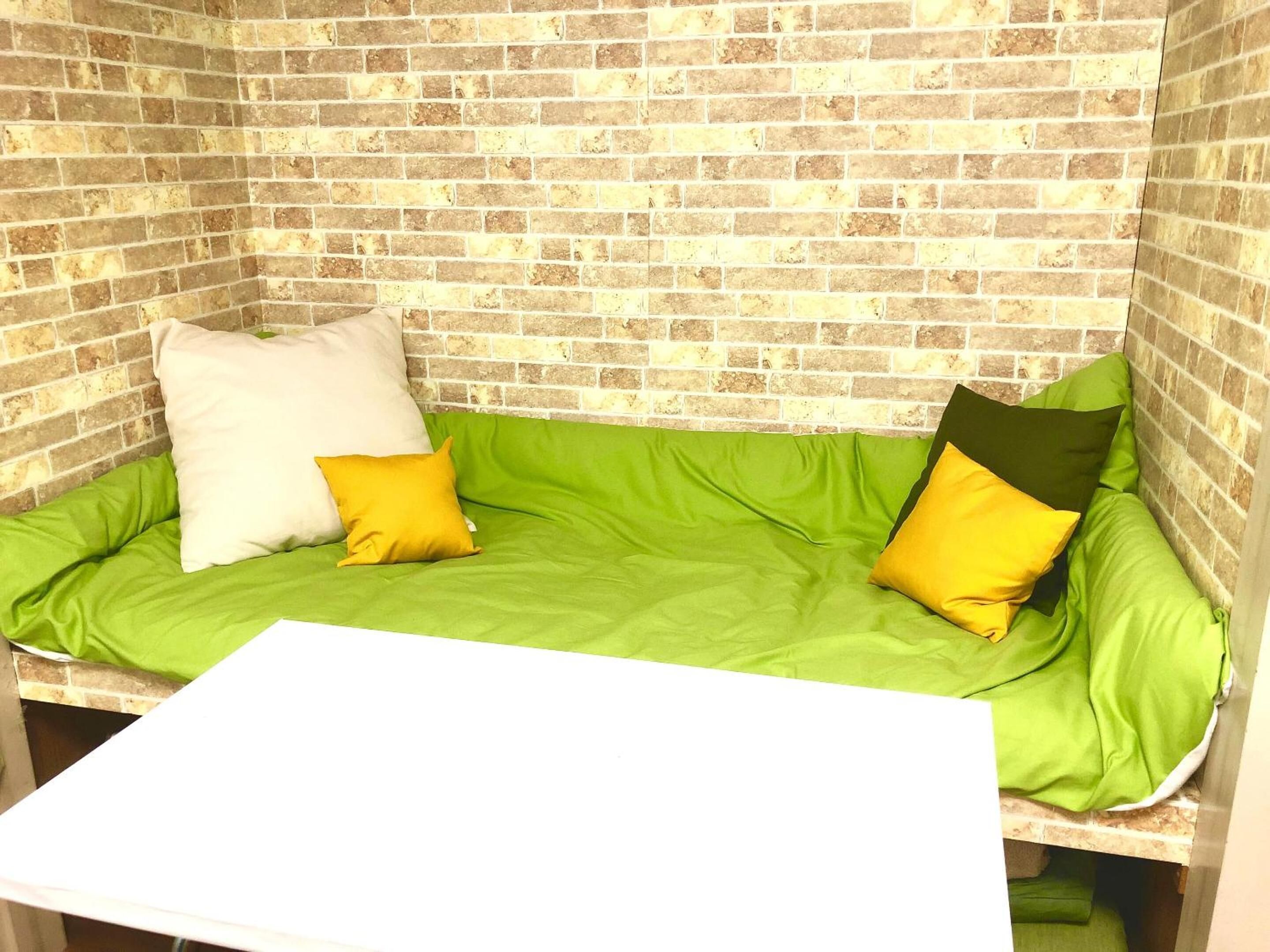 Un sofá de esquina con un cojín verde y dos almohadas, una blanca y otra amarilla, frente a una pared de ladrillo.