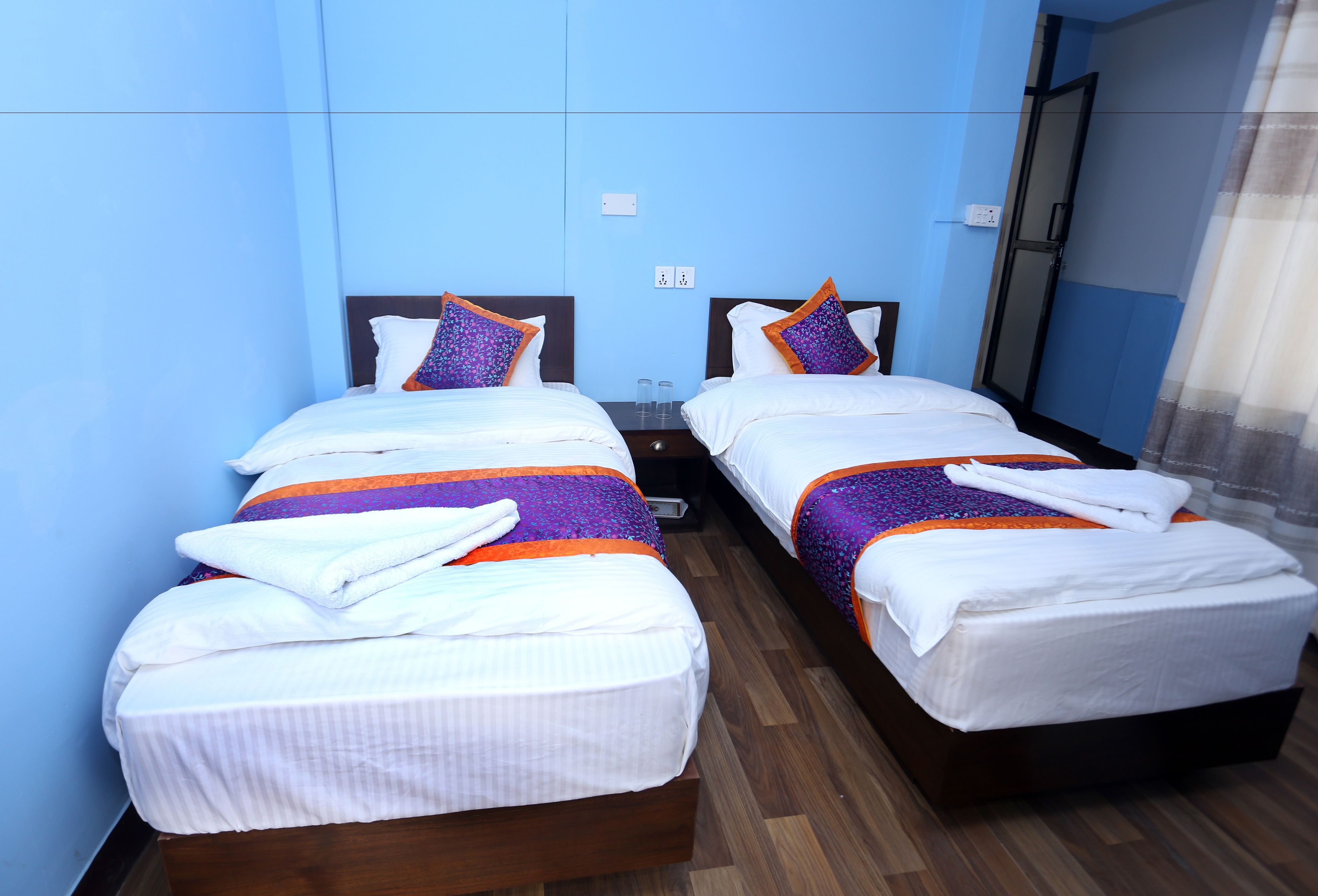 A hotel room with two beds, each with white linens and purple accents, wooden flooring, and a blue wall.