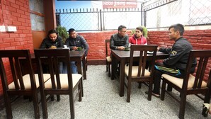 Un grupo de personas sentadas en mesas de madera con sillas, en un área de comedor al aire libre con un muro de ladrillo rojo y una cerca de metal al fondo.