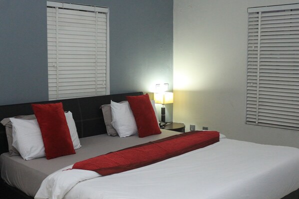 Habitación de hotel con cama, ropa de cama blanca, almohadas rojas, mesita de noche con lámpara y dos ventanas con persianas.