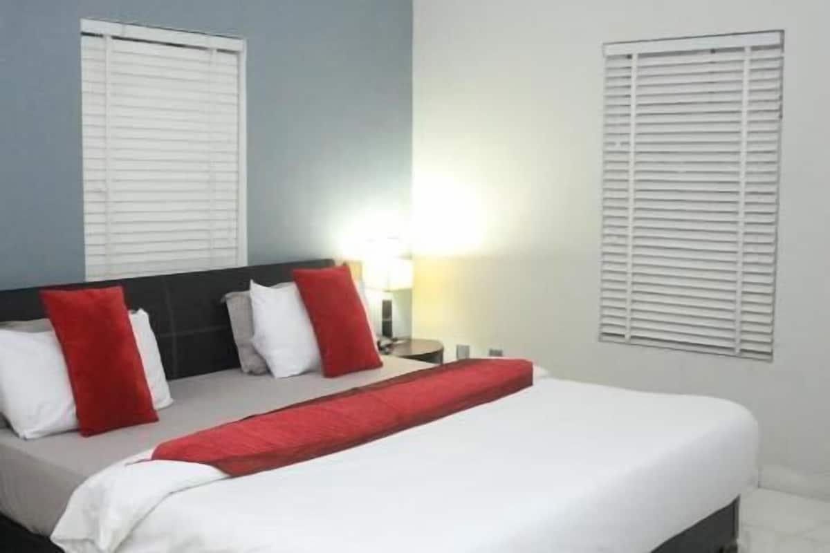 Une chambre d’hôtel avec un lit, des oreillers blancs et rouges, une table de chevet avec une lampe et une fenêtre avec des stores.