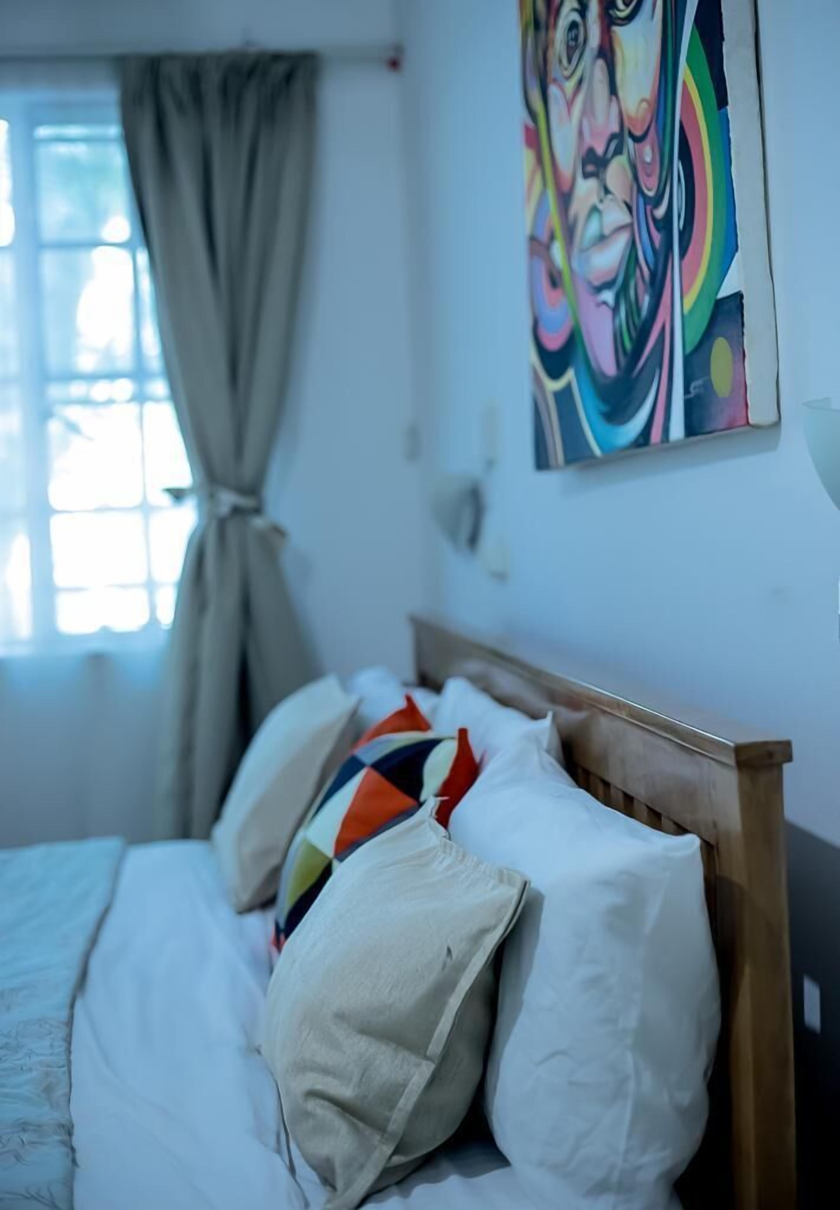 Hotellihuone, jossa on sänky, ikkuna, jossa on verhot, ja värikäs taulu seinällä.