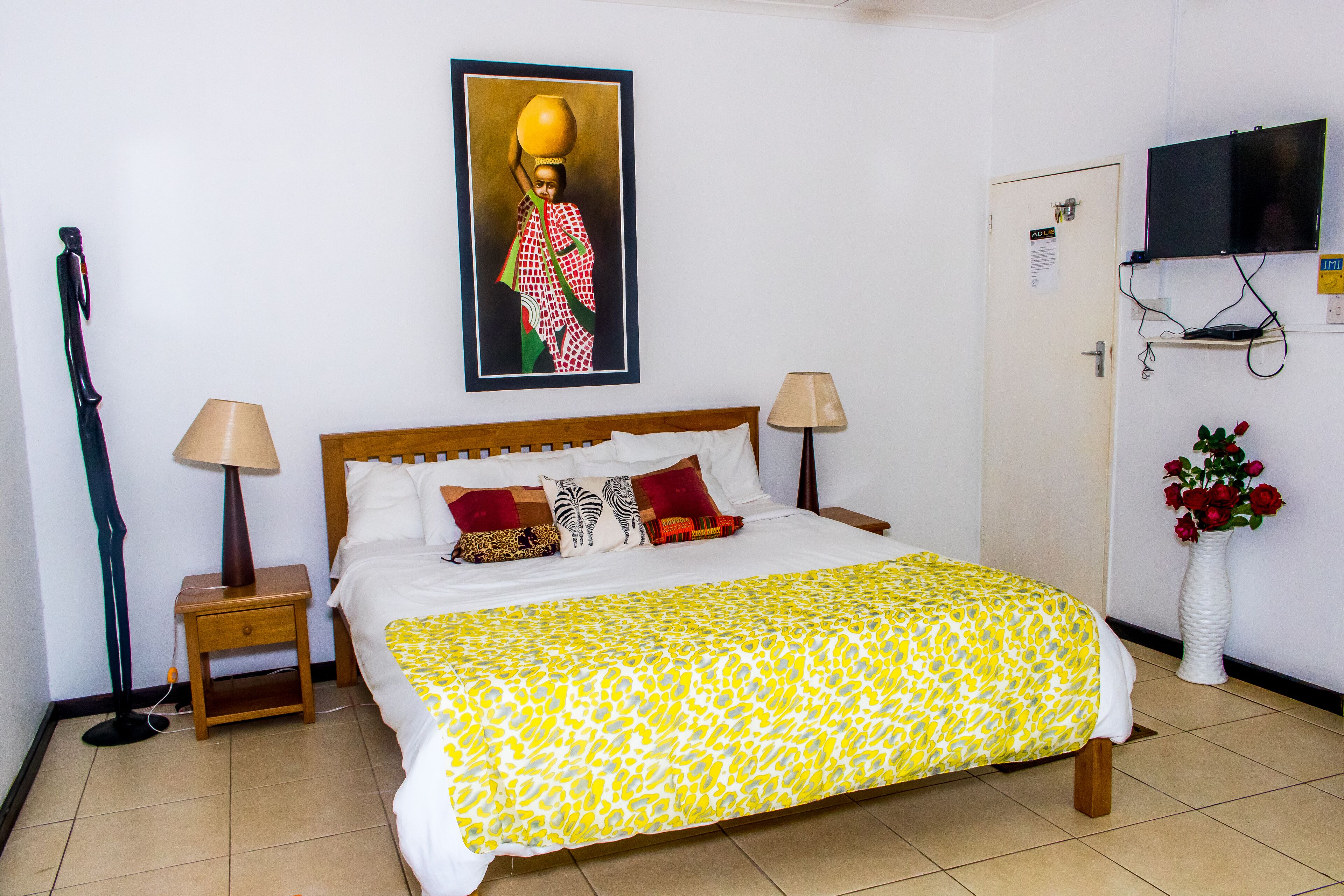 Una cama bien hecha con una colcha estampada, dos lámparas de noche, un cuadro en la pared, un televisor montado sobre una puerta, y un jarrón con flores en el suelo.