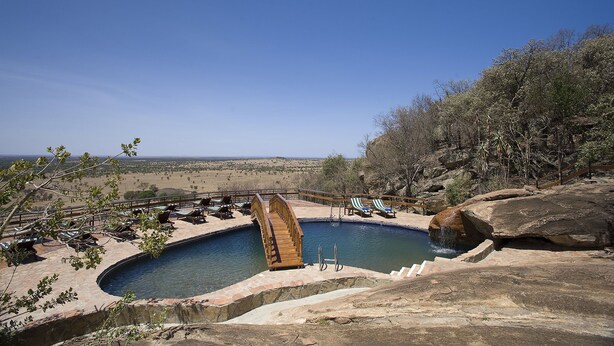 Una piscina con un puente de madera, sillas de descanso y vistas del paisaje.