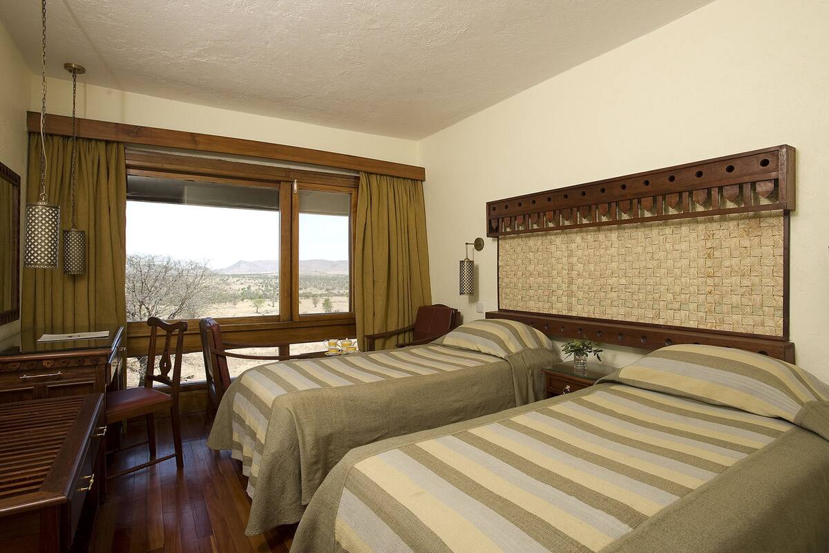 A room with two beds, a desk, and a chair. There is a window with curtains, a lamp, and a decorative headboard.