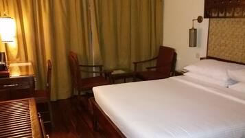 Una habitación de hotel con cama, dos sillas, un escritorio, una lámpara y una ventana con cortinas.