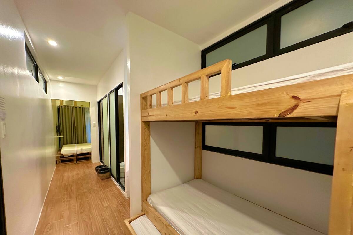 A narrow hallway with wooden flooring, a bunk bed with a mattress, a bedside table, and a window with a curtain.