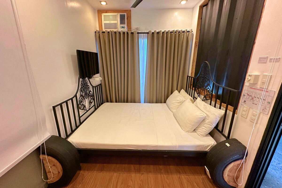 A compact, modern room with a single bed, a ceiling fan, an air conditioning unit, and a window with curtains.