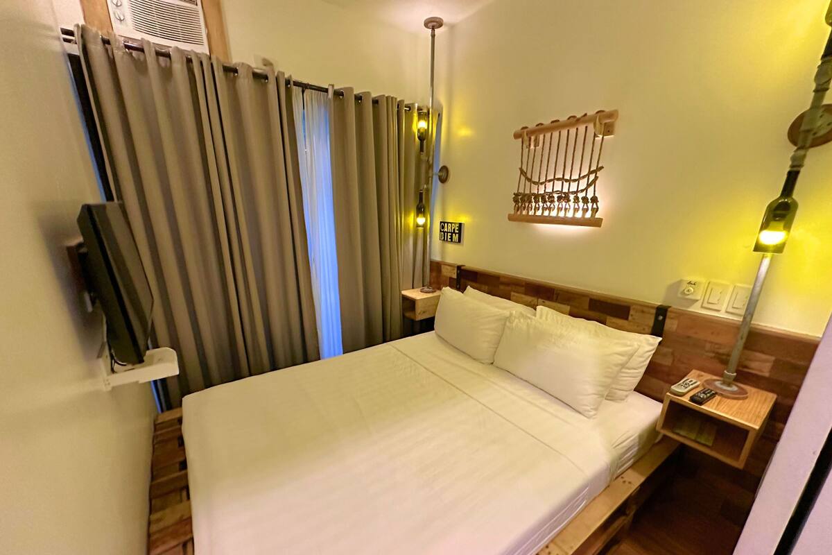A hotel room with a large bed, a wooden headboard, a nightstand with a lamp, and a wall-mounted light fixture.