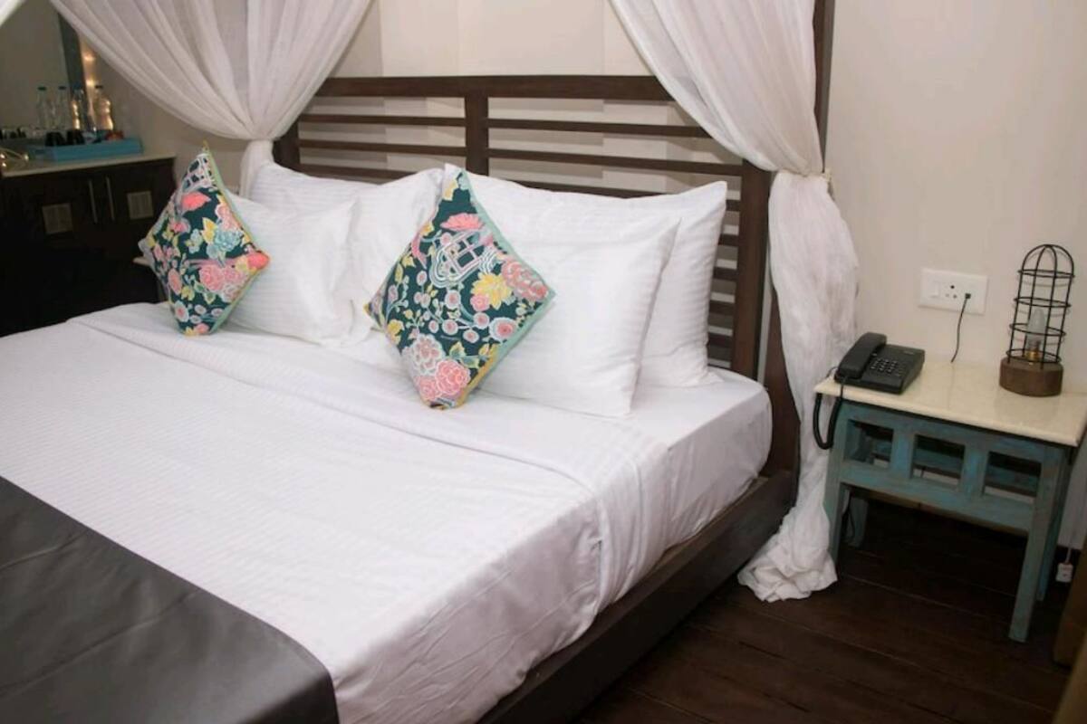 Un lit avec du linge de lit blanc et deux coussins à motifs floraux, une table de chevet avec un téléphone et un objet décoratif, et une tête de lit en bois.