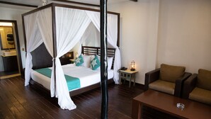 Un lit à baldaquin avec des rideaux blancs, une tête de lit en bois et une literie de couleur turquoise. On y trouve une petite table de chevet avec une lampe, un canapé marron et une table basse en bois.