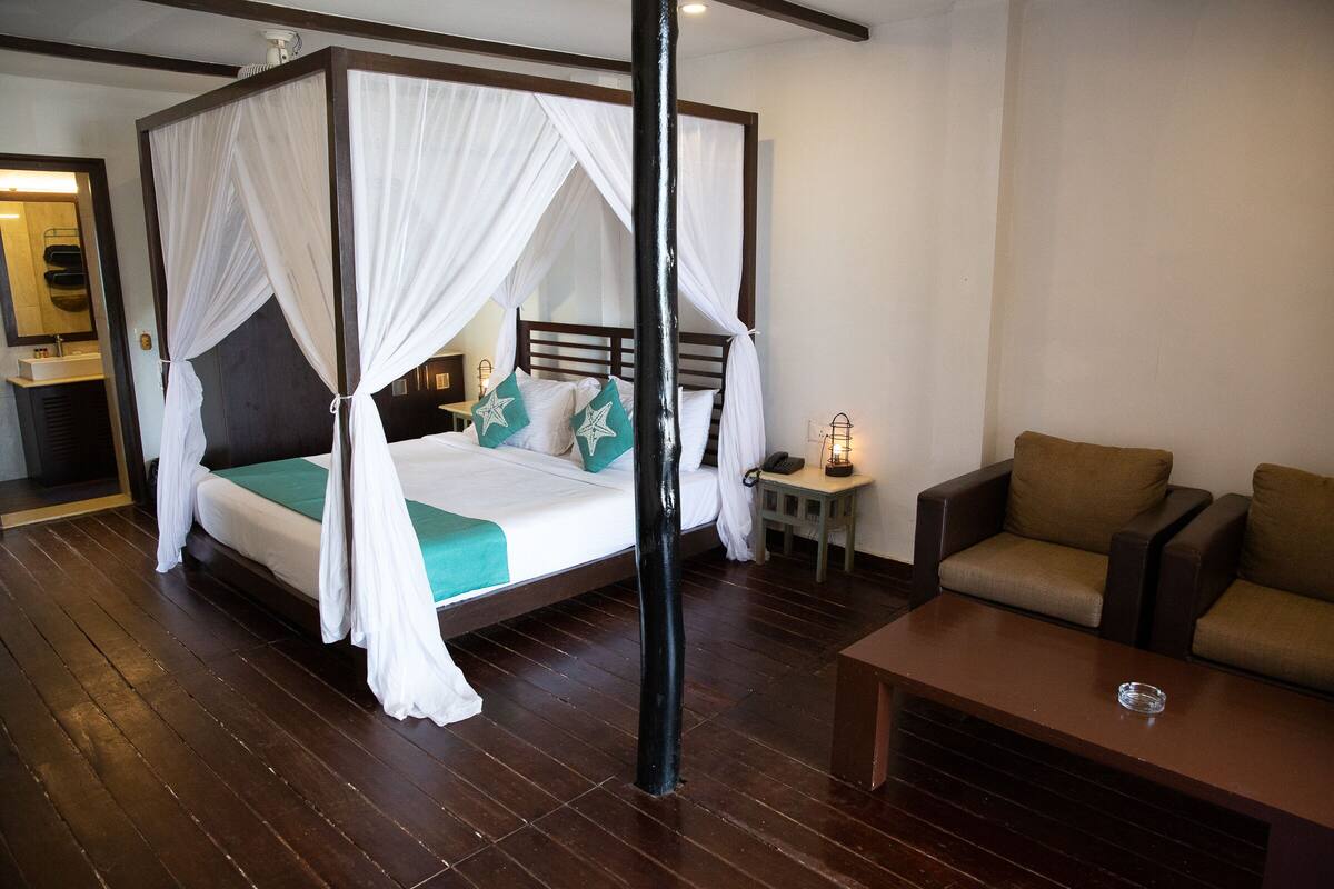 Un lit à baldaquin avec des rideaux blancs, une tête de lit en bois et une literie de couleur turquoise. On y trouve une petite table de chevet avec une lampe, un canapé marron et une table basse en bois.