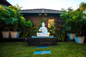 A statue of a seated figure in a garden setting with a wooden building in the background. - SinQ Beach Morjim (Morjim)