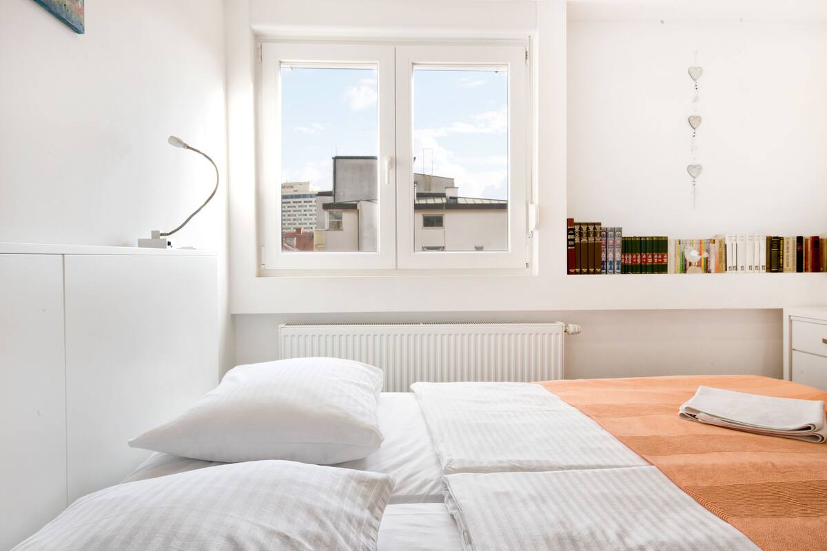 Ein Schlafzimmer mit Bett, Kissen, einem Fenster, einem Heizkörper, einem Schreibtisch und einem Bücherregal.