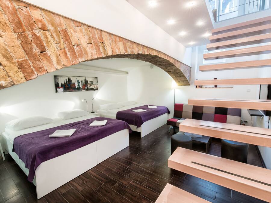 Quarto de hotel moderno com duas camas, teto em arco de pedra, escada e vista para a cidade.