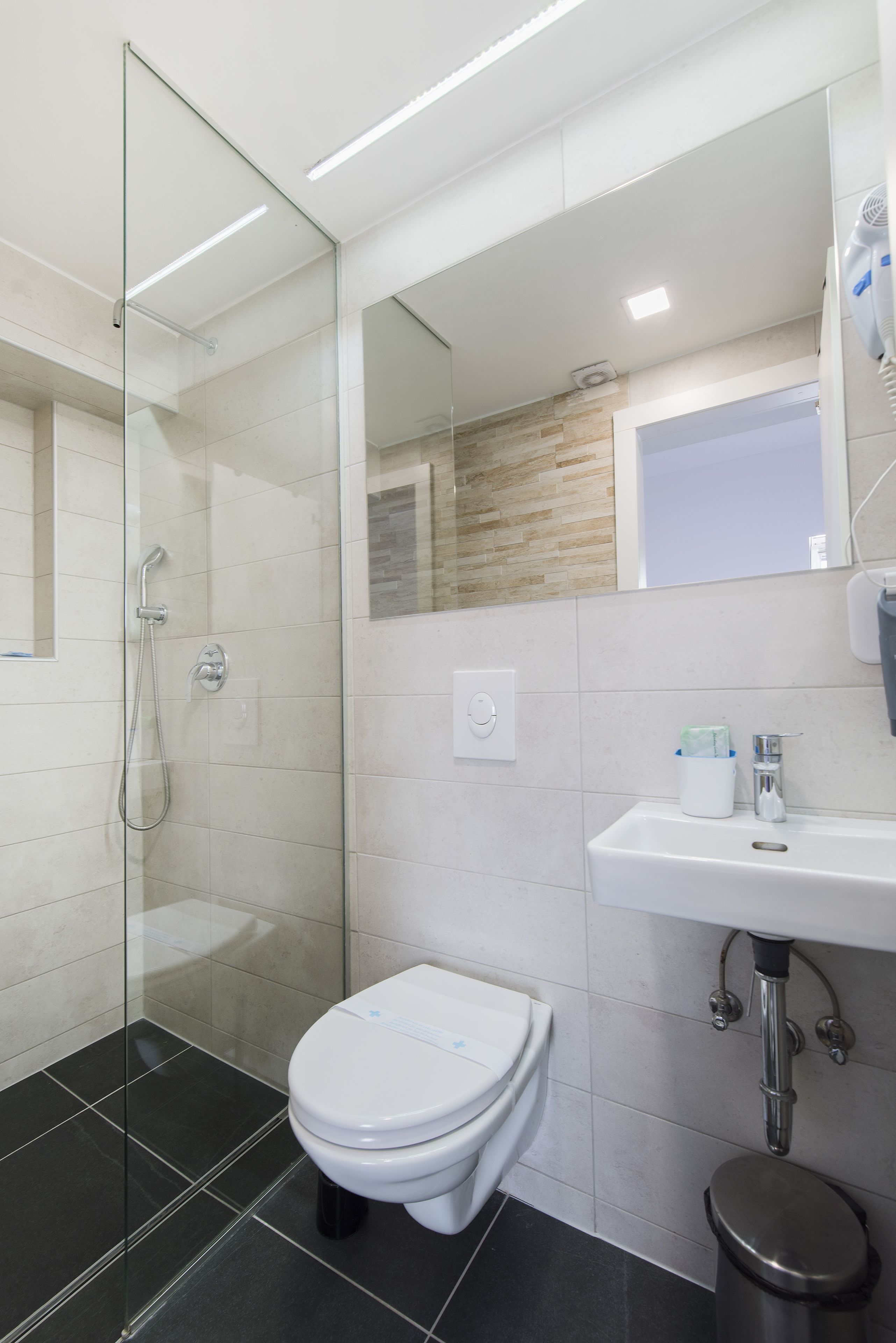 Une salle de bain moderne avec une douche à parois de verre, des toilettes blanches, un lavabo avec un miroir au-dessus et un sol carrelé.