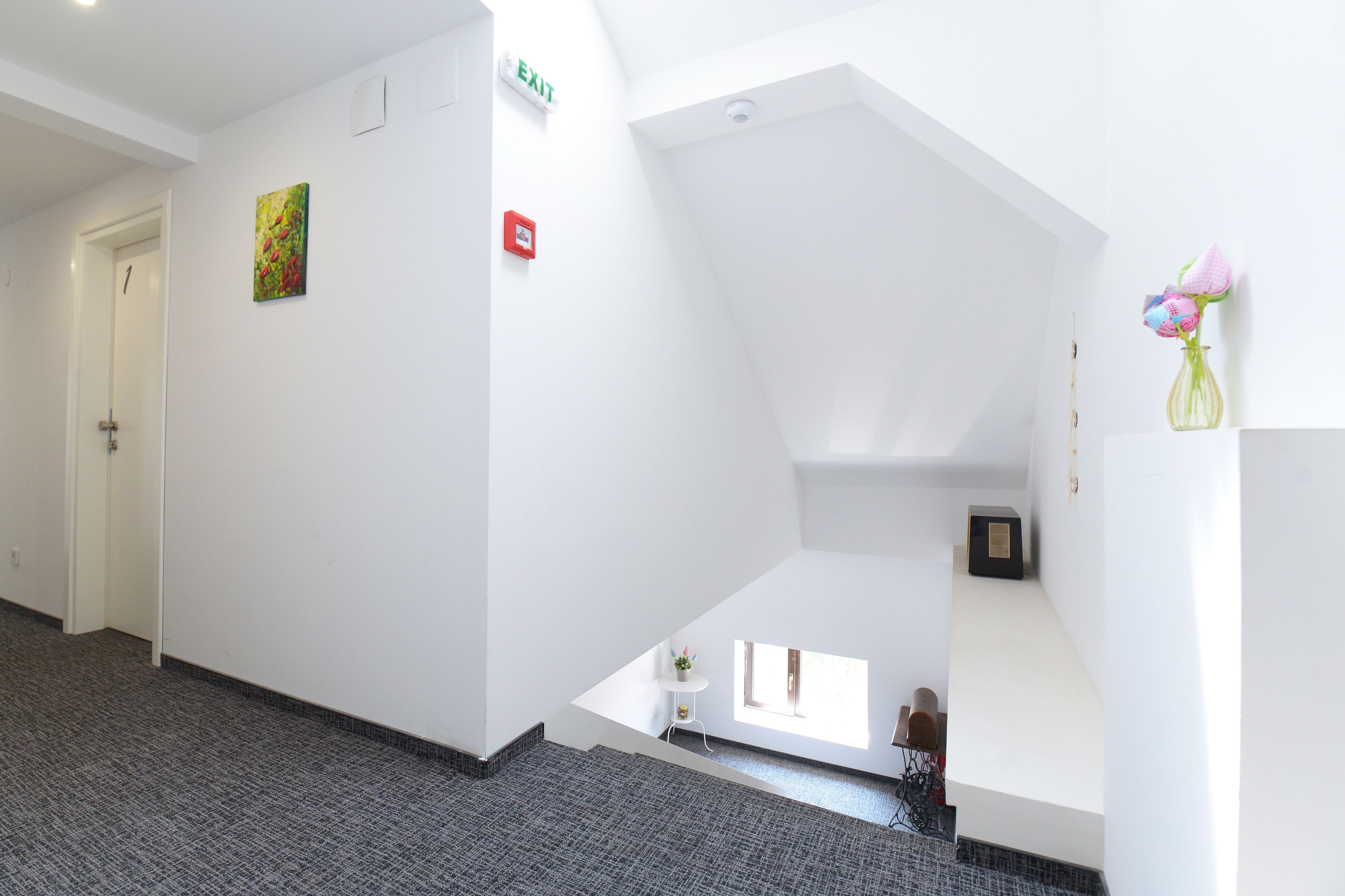 A hallway with a carpeted floor, a white door, an exit sign, a small table with a vase, and a window with natural light.