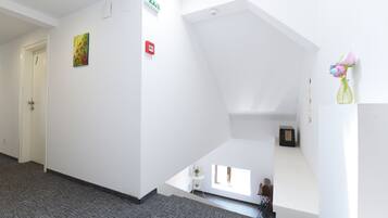 A hallway with a carpeted floor, a white door, an exit sign, a small table with a vase, and a window with natural light.