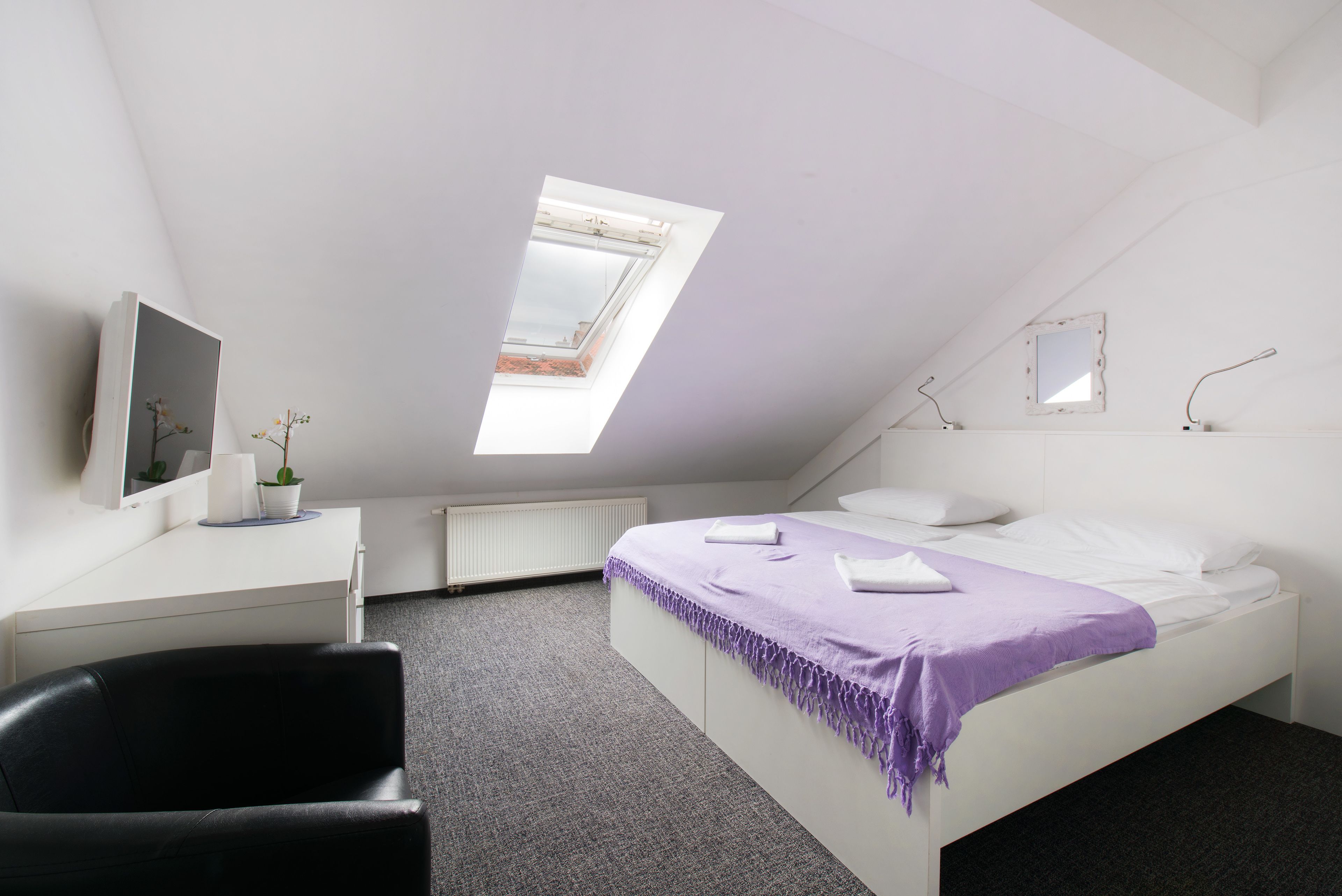 Una camera da letto con soffitto spiovente, una finestra a lucernario, un letto con coperta viola, un comodino bianco, una sedia nera e una piccola pianta.