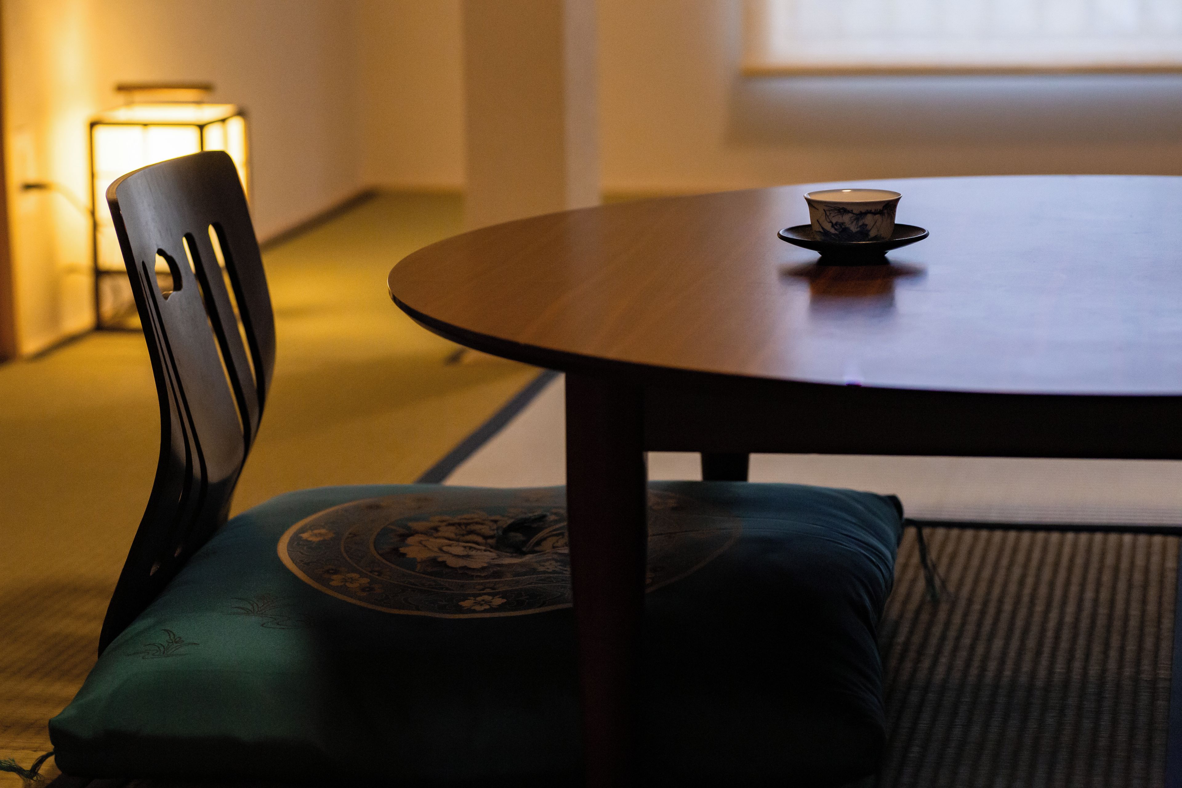 A round wooden table with a small teacup on it, a dark green upholstered chair with a floral pattern, and a lamp in the background.