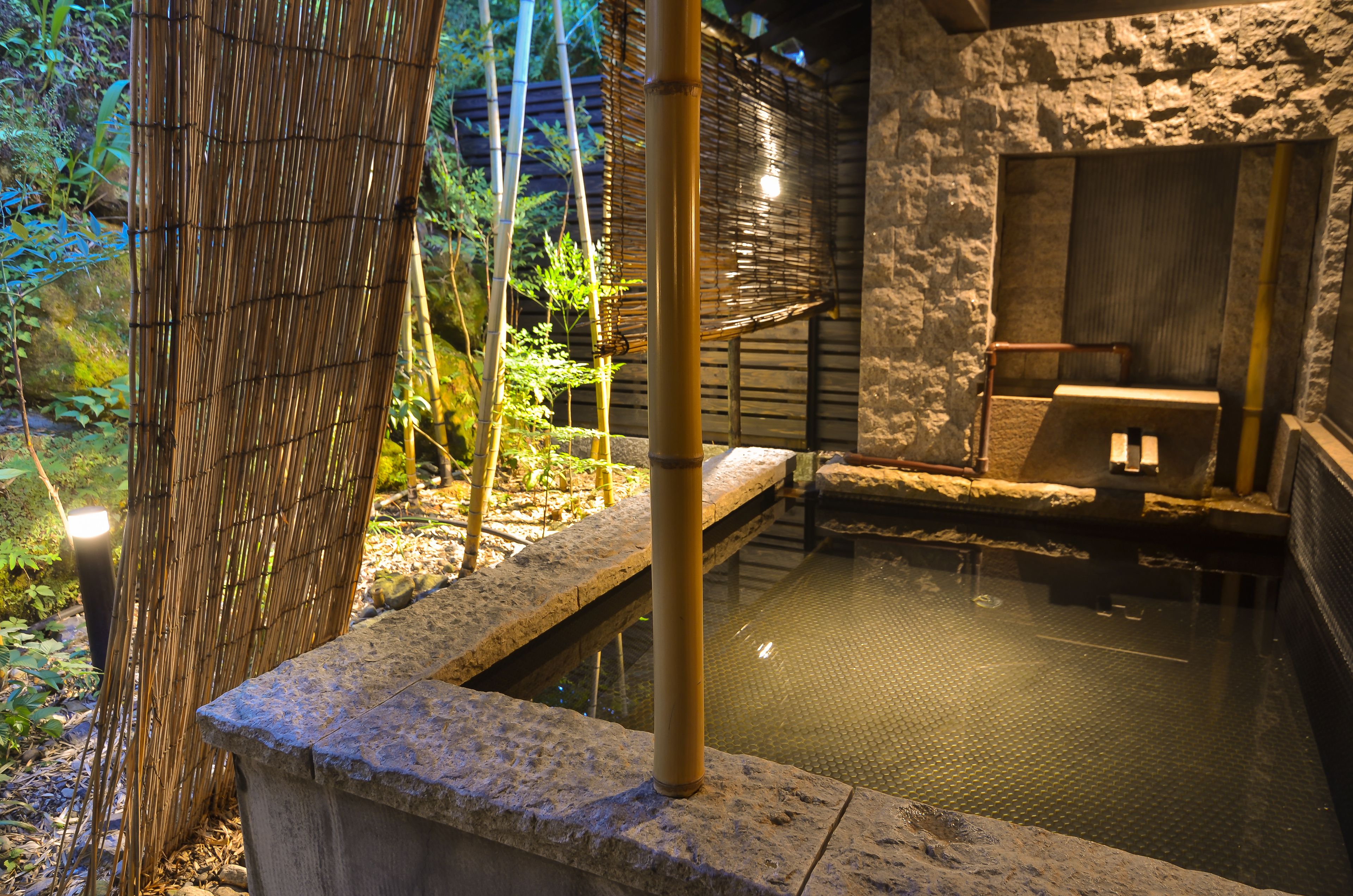 Un onsen japonés tradicional con escalones de piedra que conducen a una fuente termal, rodeado de bambú y follaje.