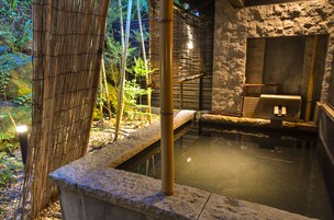 Un onsen japonés tradicional con escalones de piedra que conducen a una fuente termal, rodeado de bambú y follaje.