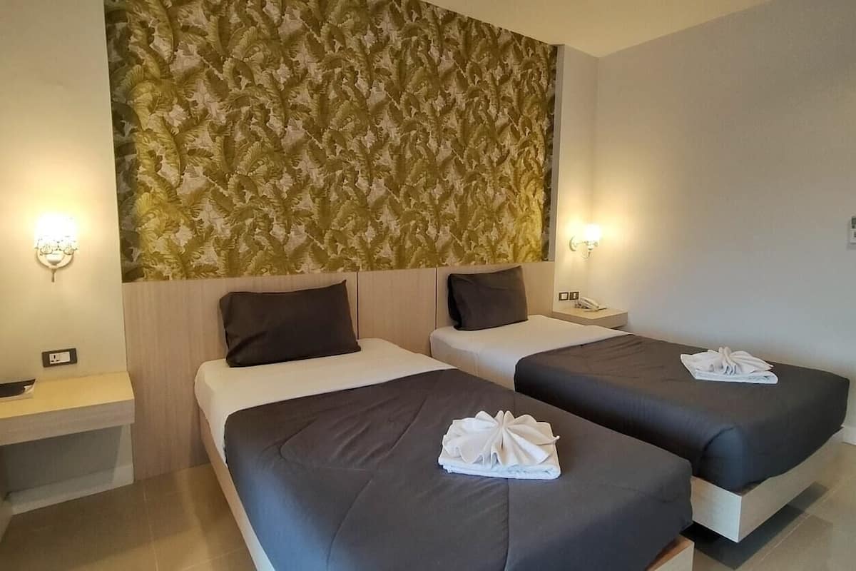 A hotel room with two beds, each with a dark-colored bedspread and a white towel on the bed. There are two bedside tables, one on each side of the beds. The room has a patterned wallpaper on the wall behind the beds.