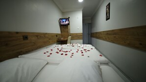 A hotel room with a wooden headboard, a bed with white linens and rose petals, a TV on the wall, and a door in the background. - Otel Altay (Samsun)