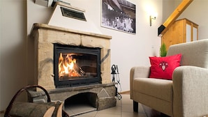 A cozy living room with a stone fireplace, a grey sofa with a red cushion, and a wicker basket with logs.