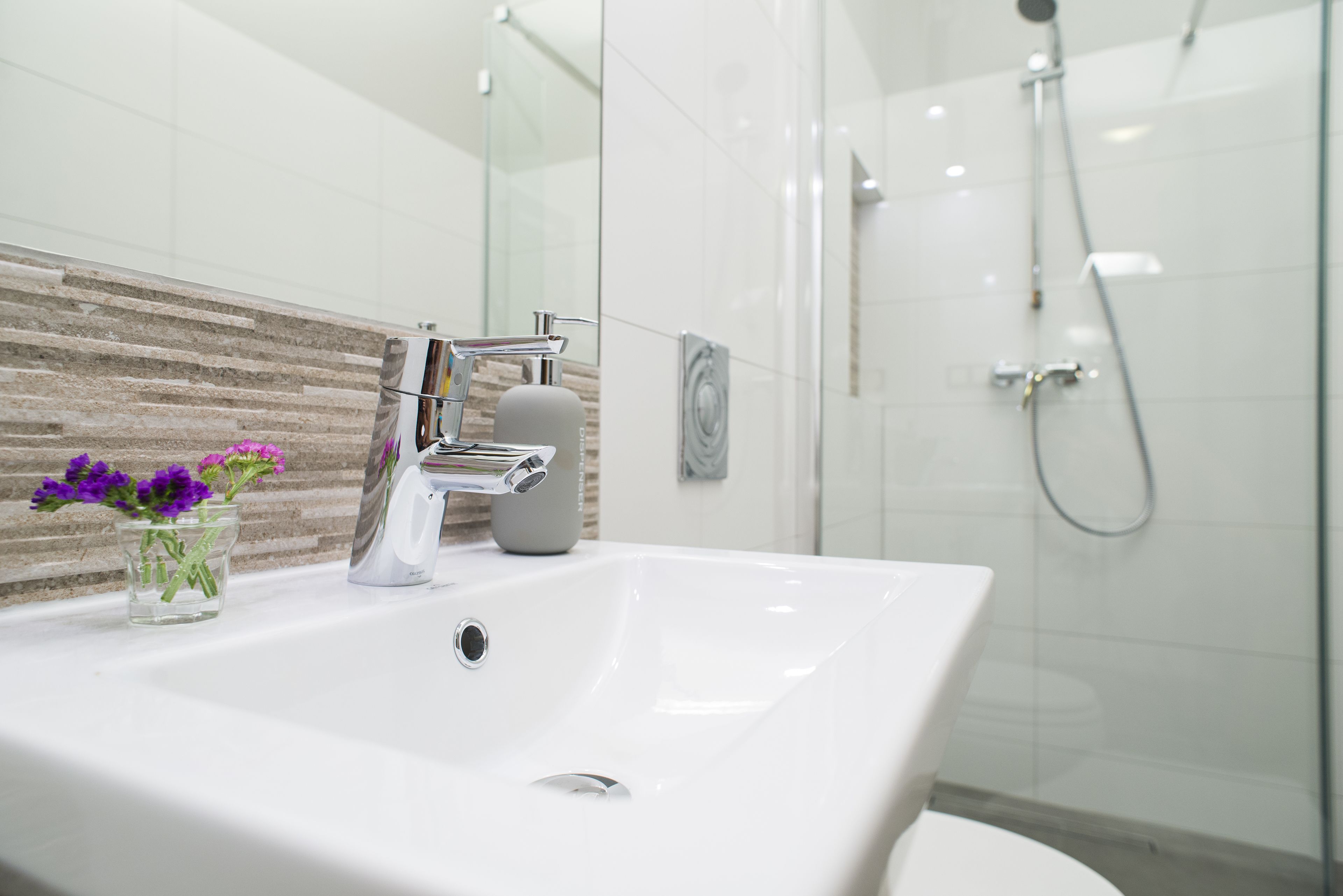 A modern bathroom with a white sink, a glass vase with purple flowers, a shower area, and a tiled wall.