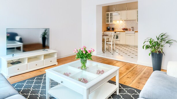 A modern living room with a flat-screen TV, a glass coffee table with flowers, a sofa, and a kitchen visible through an open doorway. -  Apartament Nowe Miasto - Park Róż (Gorzow Wielkopolski)