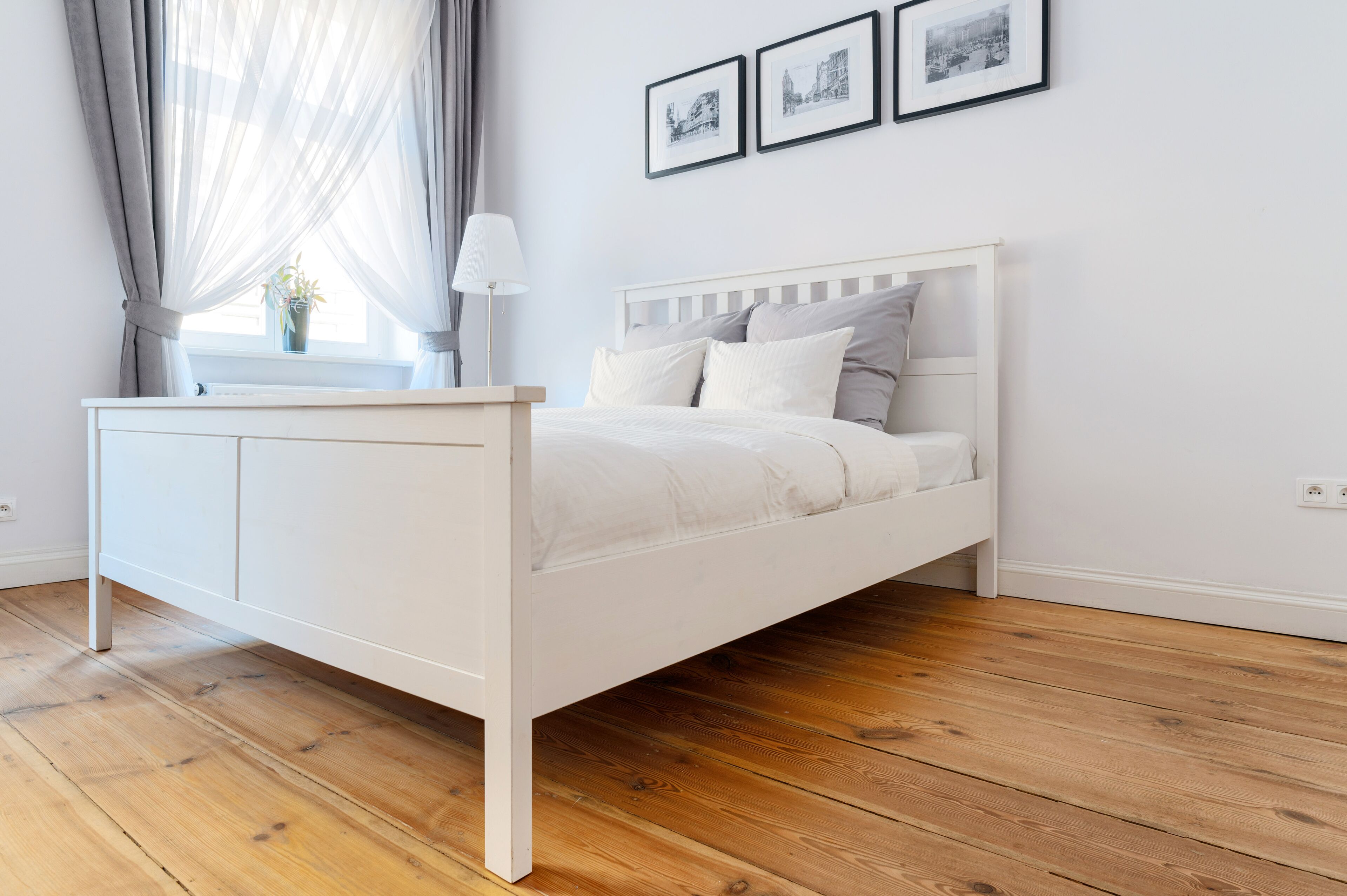 A white bed with a wooden headboard in a room with wooden flooring and a window with sheer curtains.