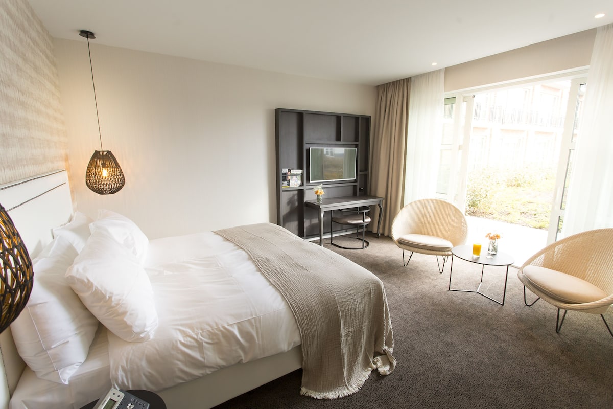 Habitación de hotel con una cama, un escritorio con televisor, dos sillas y un ventanal grande.